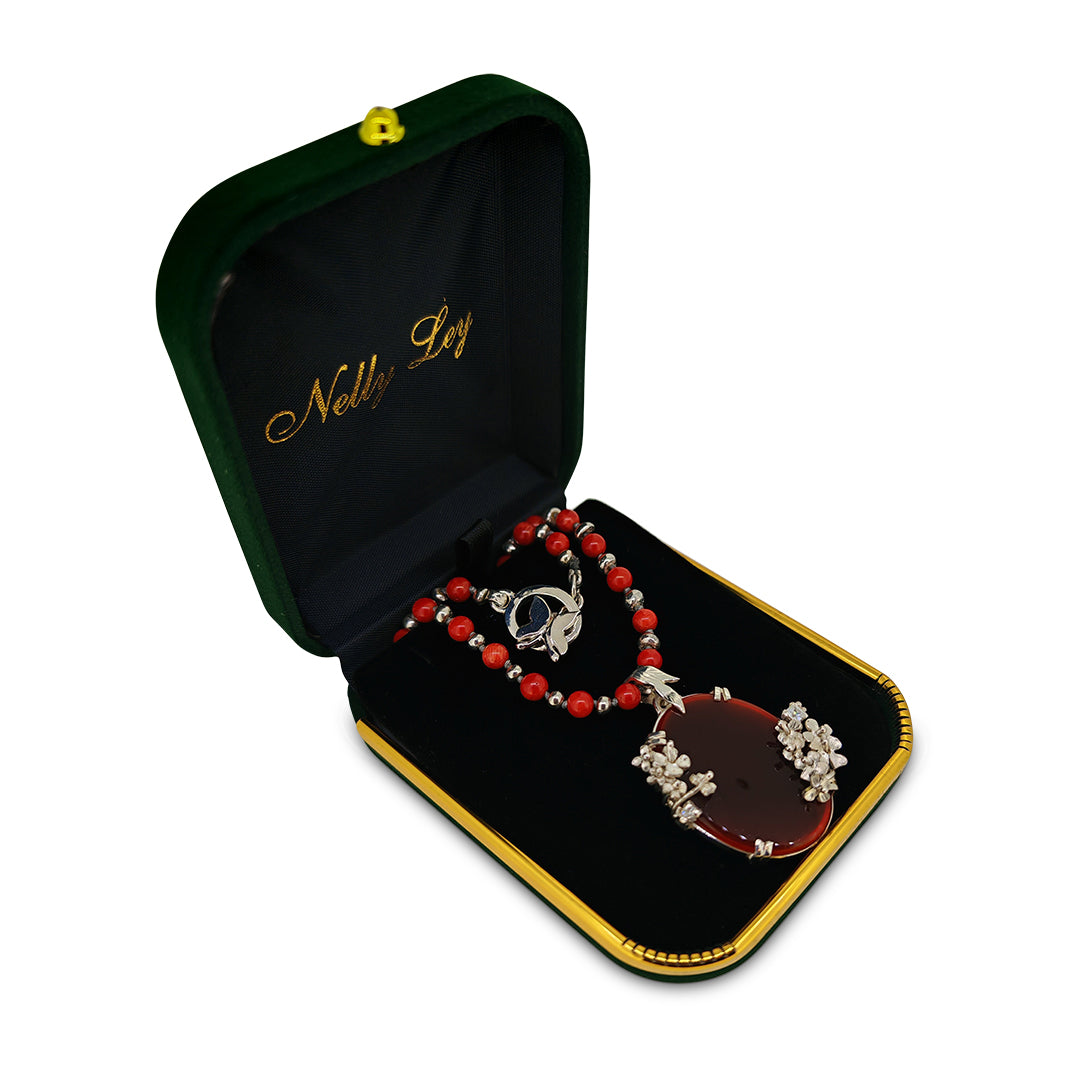 Necklace with red beads and a pendant in an open jewelry box with 'Nelly Ley' branding.